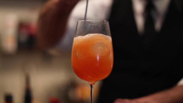 A bartender stirs an orange cocktail in a wine glass, with bubbles, condensation, and a warm blurred bar background