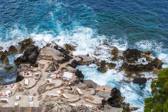 View of sunbathers lounging on rocky terraces as turquoise waves crash against the rugged coastline, Capri, Campania, Italy.