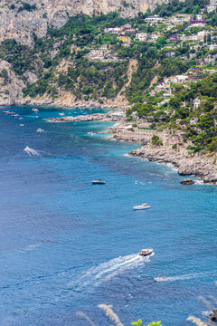 View of boats cutting through the vibrant blue Tyrrhenian Sea against the backdrop of rugged cliffs and verdant hills, Capri, Campania, Italy.