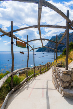 View of a winding path with wooden pergola framing the turquoise sea dotted with boats, against rugged cliffs under a bright sky, Capri, Campania, Italy.