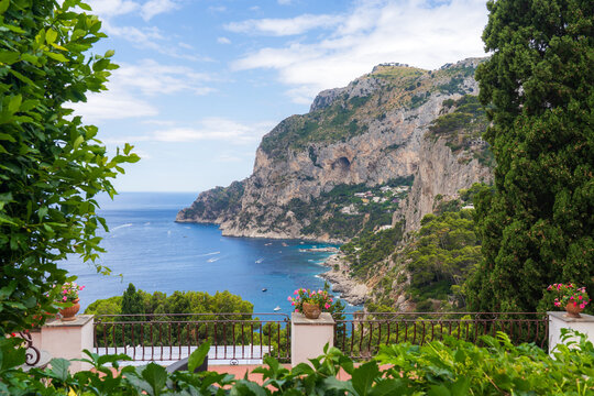 View of dramatic cliffs meet the azure sea, framed by lush greenery and ornate balustrades under a sky streaked with clouds, Capri, Campania, Italy.