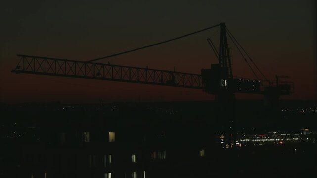 Dramatic silhouette of a large construction tower crane standing motionless against a beautiful, deep red sunset sky, with the twinkling lights of the city coming alive in the evening. timelapse