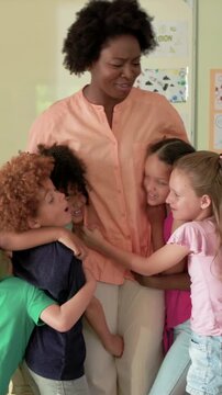 Vertical video: Children entering right hugging teacher in classroom with TV, peach blouse for hugs