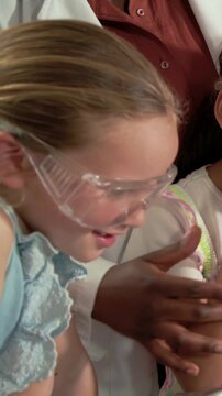 Vertical video: Picking right kid holding cup at lab, goggles, lab-coat teacher guiding, copy space