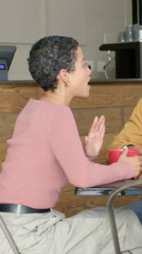 Leaning forward woman in pink ribbed sweater gesturing at cafe table, holding red mug