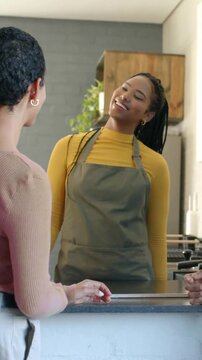 Staff member in mustard top, gray apron arriving behind counter smiling, leaning, serving customer