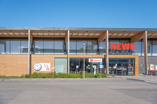 Germany Berlin April 26, 2026. Modern supermarket front with glass windows and wooden facade. Bright red sign stands out against blue sky. Clean pavement and green shrubs line the entrance. No people 