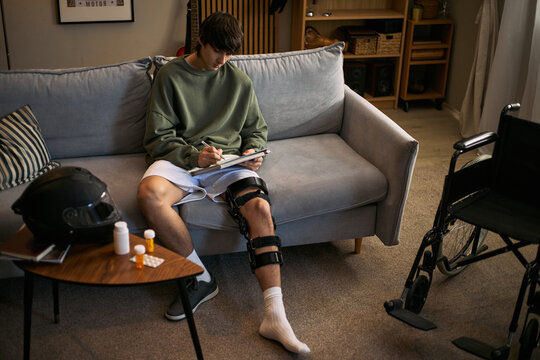 Teenager boy wearing knee brace, writing notes on sofa beside wheelchair and medicine after injury recovery. Useful for rehabilitation, home care, insurance, mobility support marketing
