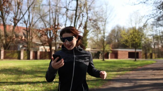 Runner woman checks smartphone while jogging in park distracted from workout losing focus on training pace digital addiction habit active lifestyle struggle outdoor fitness