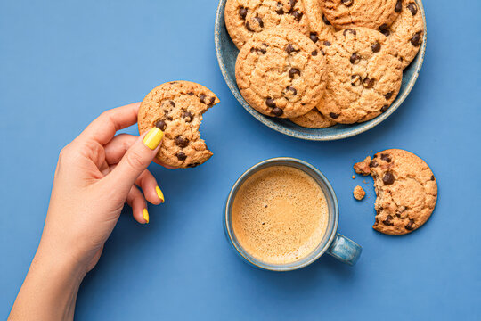 A hand holding a bitten cookie with a cup of coffee and a plate of cookies on a blue background.
