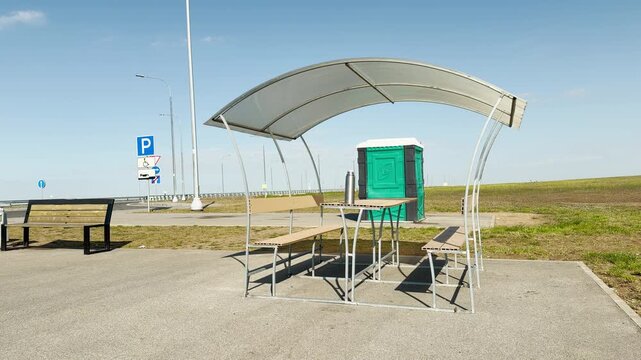 A vibrant green portable toilet sits near a modern covered picnic bench under a clear blue sky. This quiet rest area offers a peaceful, convenient break for weary travelers on the road.