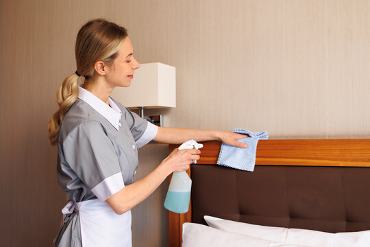 Professional housekeeper dusting hotel bed headboard with cloth and cleaning spray