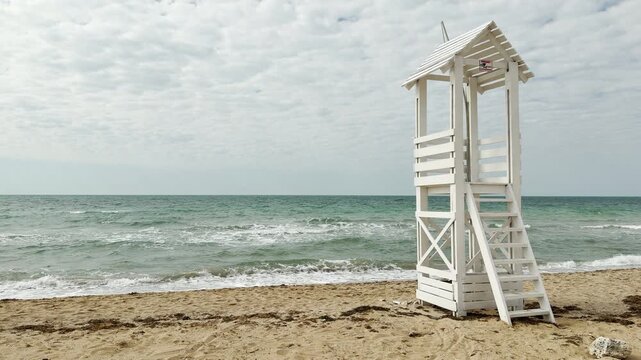 A wooden lifeguard station stands lonely on a quiet beach under a cloudy sky. The turquoise waves and empty sand create a peaceful, slightly lonely feeling of a season ending.