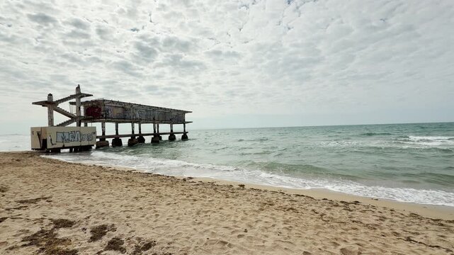 A weathered concrete pier covered in graffiti stretches into the calm Black Sea. The decaying structure evokes a sense of haunting nostalgia against the bright, sandy beach and cloudy sky.