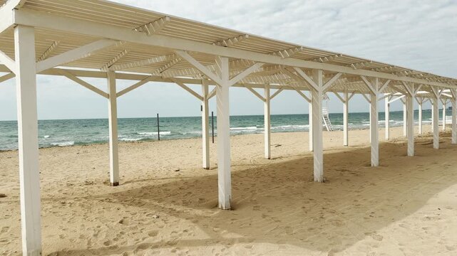 Long white wooden shelters stretch across the empty sandy beach. The clean lines of the structures create a peaceful, orderly feeling against the blue sea. This quiet scene feels calm and inviting