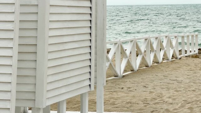 A clean white wooden cabin and fence sit quietly on the sandy beach. The cool blue waves create a calm and refreshing atmosphere. This peaceful scene feels light, airy, and very inviting.