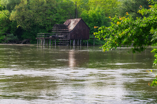 View of a rustic, weathered wooden mill house standing on stilts in the tranquil, shimmering waters, embraced by lush green trees, Tomasikovo, Trnava Region, Slovakia.