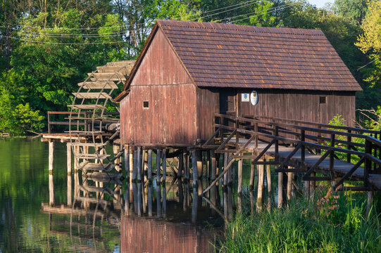 View of a rustic wooden watermill house standing above a tranquil lake, its reflection shimmering in the water, connected by a wooden bridge, Tomasikovo, Slovakia.