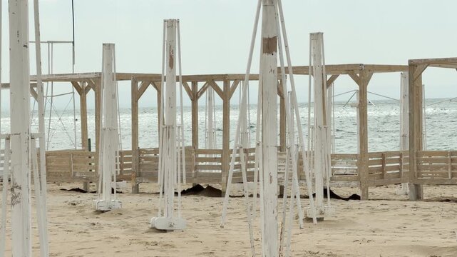 Stripped white umbrella frames stand lonely on the quiet sand. The wooden gazebos behind them face a shimmering sea, creating a melancholic yet peaceful mood. This scene feels still and nostalgic.
