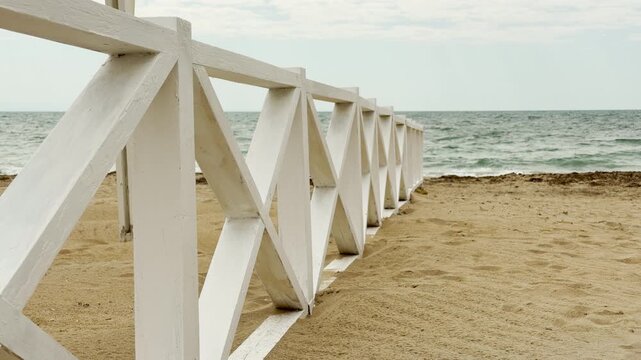A geometric white wooden fence stretches across the soft sand toward the water. The overcast sky creates a quiet and thoughtful mood. This peaceful coastal scene feels still and refreshing.