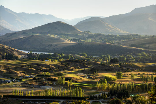 View of a hilly landscape with golden sunlight illuminating the green and brown fields, a river snaking through the hills, , Arrowtown, Otago Region, New Zealand.