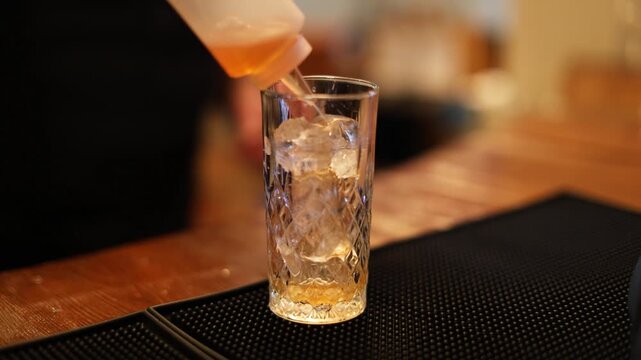 A bartender pours clear spirit into an ice-filled glass at a bar, beginning an orange cocktail that is later finished with juice and stirring