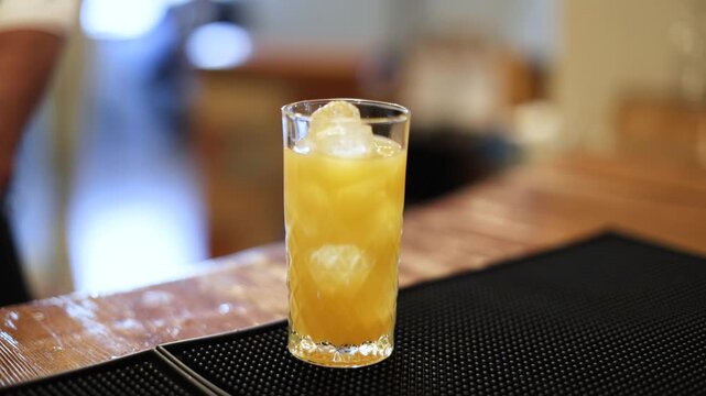 A bartender finishes a layered red and orange cocktail with ice, dried citrus, and a yellow flower garnish in a tall glass against a warm blurred bar background