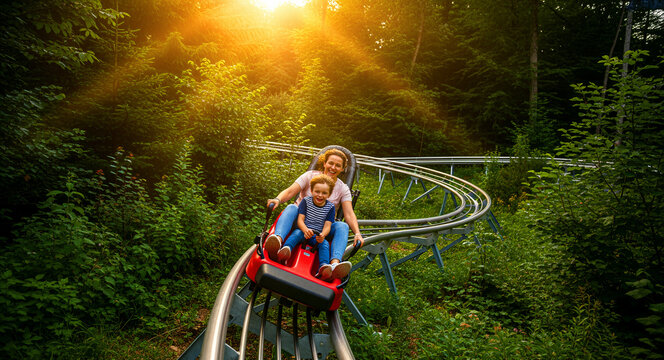 Happy mother and son riding a mountain coaster through a lush forest. The family enjoys a thrilling ride on an alpine slide during a sunny day. Summer outdoor activity and tourism concept.