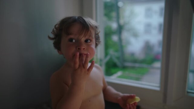Toddler eating apple snack by window while looking at camera, curious expression and quiet moment of childhood in calm home environment