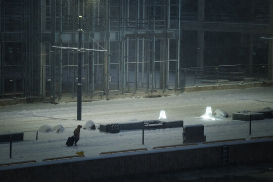 View of a solitary figure trudging through the snowy cityscape with luggage, under the pale glow of streetlights, in the quiet stillness of winter, Tallinn, Estonia.