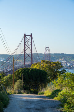 View of a dirt road leading to a striking red bridge spanning the Tagus River, framed by vibrant green foliage under a clear blue sky, Lisbon, Lisbon, Portugal.