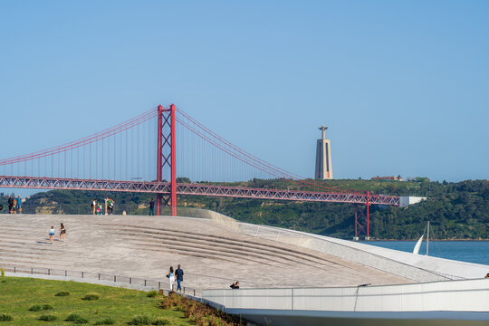 Lisbon, Portugal - 29 April 2022: View of the iconic red 25 de Abril Bridge and majestic Cristo Rei statue, contrasting with modern concrete terraces and serene blue waters.