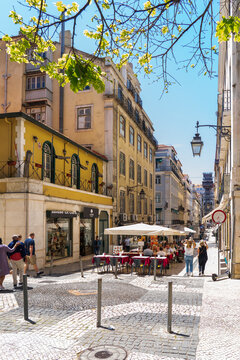 Lisbon, Portugal - 08 May 2022: View of a charming, sun-drenched cobblestone street lined with historic yellow buildings and a vibrant outdoor cafe under a clear blue sky.