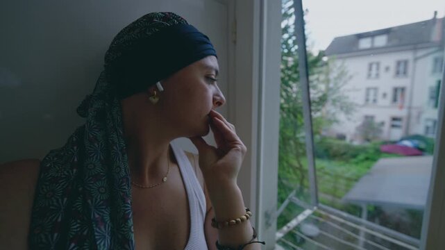 Cancer treatment isolation and introspection, woman sits on window ledge looking outside, withdrawn posture expressing emotional burden and quiet endurance in recovery process