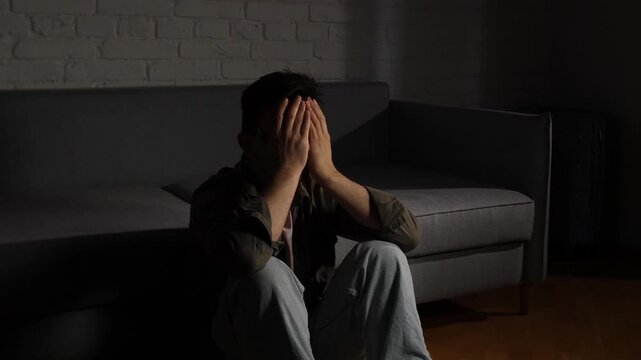 Man covering his face with hands, sitting on the floor in a dark room, experiencing sadness, depression, and loneliness in a moment of emotional crisis