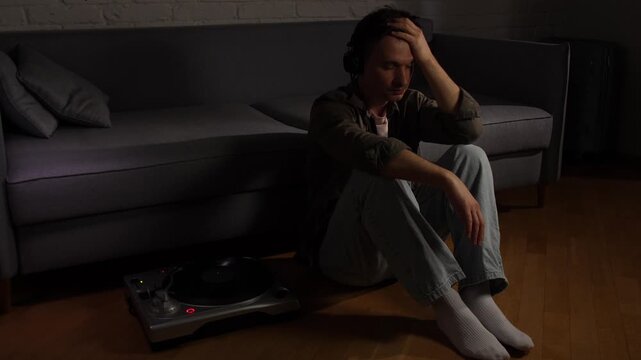 Young man seated on a dim floor, headphones on, listening to vinyl in silence, lost in melancholy and exhaustion, quiet solitude and emotional struggle in a dark room
