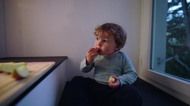 Toddler eating apple snack close up, expressive face and hand to mouth gesture showing curiosity, taste exploration and natural feeding behavior in home setting