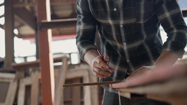 Skilled male carpenter using a pencil and square ruler to mark a wooden plank in a workshop. Professional craftsman measuring timber for a woodworking project in a rustic industrial studio
