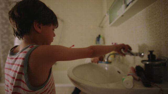 Older child turns on faucet while brushing teeth as toddler imitates in background, candid sibling interaction showing learning, curiosity and shared routine in bathroom