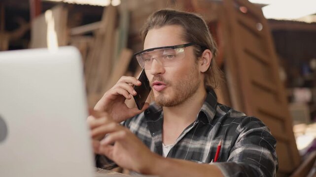 Busy male carpenter talking on a smartphone while using a laptop in his woodshop, focused on multitasking, client communication, and managing orders for his professional furniture business