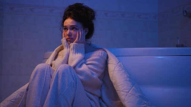 Young, sad woman wrapped in a blanket sitting on the bathroom floor, feeling desperate and upset. A lonely, heartbroken female dealing with mental health