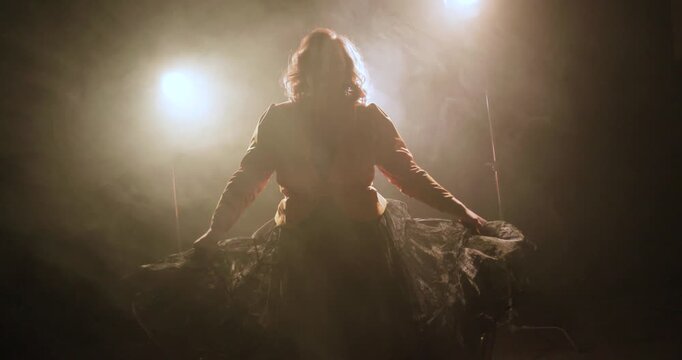 A dancer performs in slow motion on a dark smoky stage, lifting a flowing skirt under bright backlights for a dramatic theatrical silhouette scene