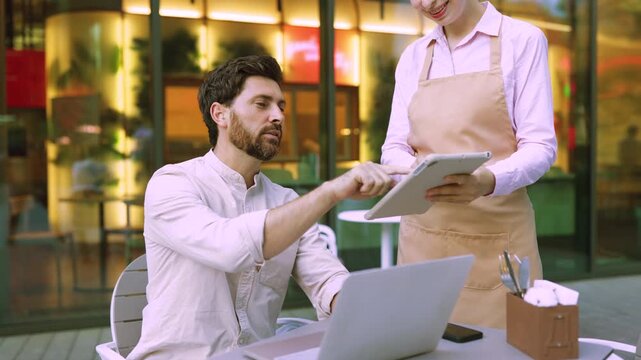 A man points at a tablet held by a server while sitting at an outdoor cafe table with a laptop