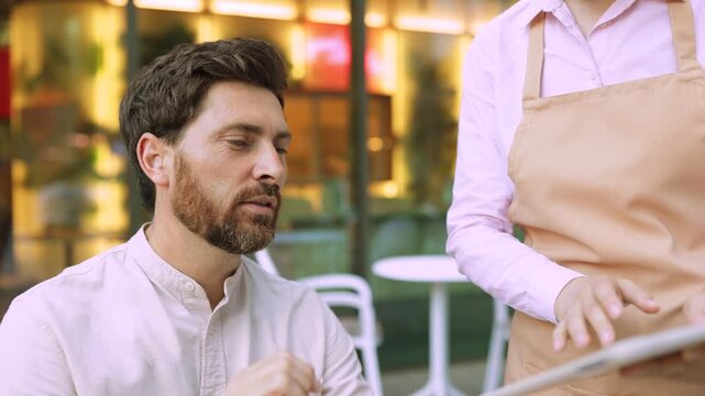 A man points at a tablet screen while a server wearing an apron takes his order at an outdoor cafe