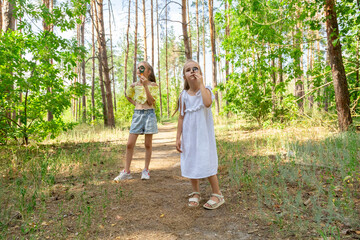 Fototapeta premium Two kids in casual clothes in summer exploring nature in forest together. Little girl looking through magnifying glass while squatting on ground