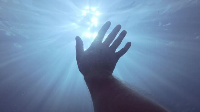 POV underwater video: Morning dancing sunbeams pierce surface of water, man's hand waves in greeting to new day under water, silhouette of bottlenose dolphin swims overhead, backlit by sun rays.