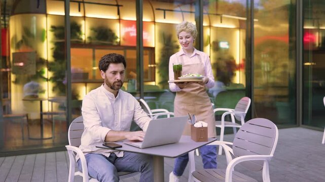 A smiling waitress in an apron brings a tray with a salad and a drink to a man working on his laptop at an outdoor cafe table