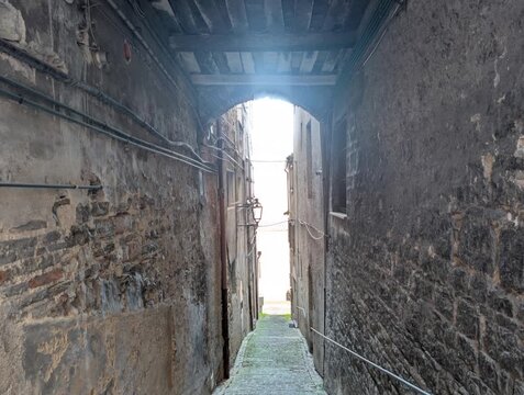 Vicolo stretto e ombreggiato tra edifici in pietra, con travi in legno e uscita luminosa in fondo all&rsquo;arco. Una scena dal forte carattere storico che evoca atmosfera antica