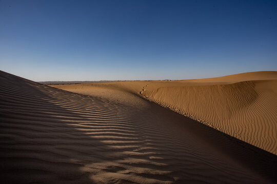 View of golden sand dunes stretching endlessly under a brilliant blue sky, with deep shadows highlighting rippling textures, Bahawalpur, Pakistan.