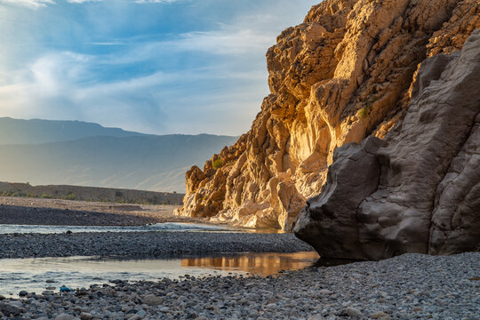 View of the sun-kissed cliffs casting long shadows over the rocky riverbed, contrasting with the cool blue waters, Pakistan.